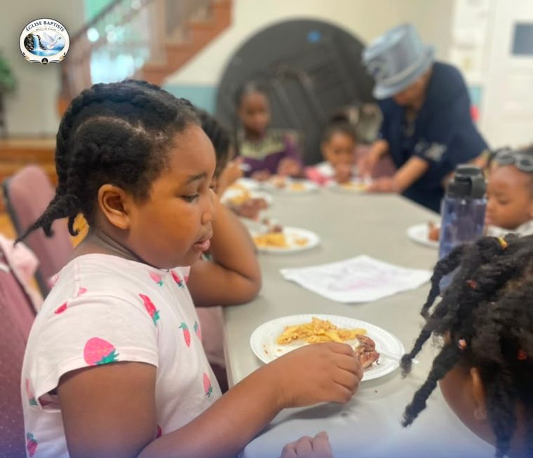 Une fillette mange à table ; derrière elle, d'autres enfants et une dame en chapeau qui les sert.